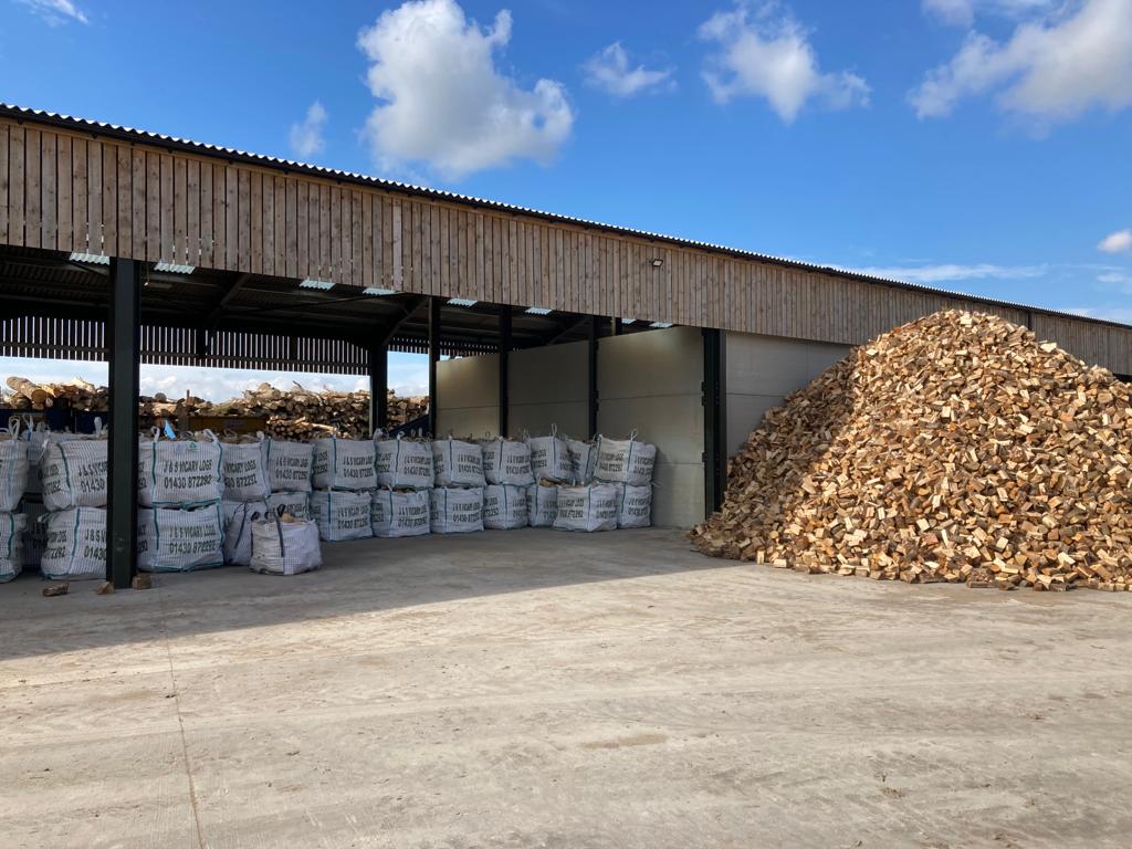 Storage sheds stocked with bagged firewood beside a stacked pile of logs